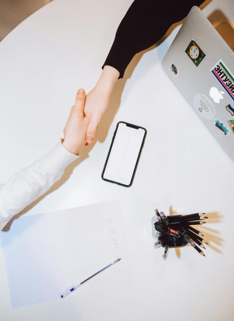 Two individuals shaking hands across a desk with a smartphone and laptop in a modern office setting.
